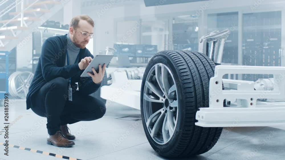 Aufkleber Engineer with Glasses and Beard Works on a Tablet Computer Next to an Electric Car Chassis Prototype with Wheels, Batteries and Engine in a High Tech Development Laboratory.