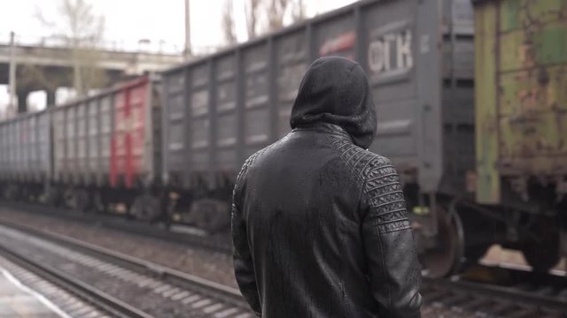 Young man looking at passing freight train. Young man in leather jacket with hood looking at passing freight train in rainy day