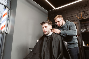 Master cuts hair and beard of men in the barbershop, hairdresser makes hairstyle for a young man. Barber work with clipper machine in barbershop.