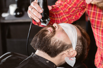 Beard cutting, face care. Barber work with clipper machine in barbershop. Professional trimmer tool cuts beard and hair of young guy in barber shop salon.