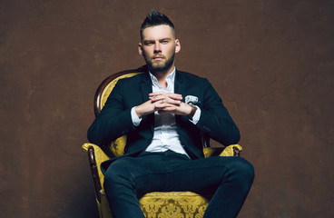 Young handsome man posing for a fashion shoot in a studio. Fashion as a lifestyle. Man wearing a beard. Model sitting on a sofa in tuxedo. Successful fashionable businessman. Business look advertising