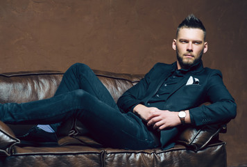 Young handsome man posing for a fashion shoot in a studio. Fashion as a lifestyle. Man wearing a beard. Model sitting on a sofa in tuxedo. Successful fashionable businessman. Business look advertising