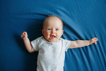Portrait of cute adorable smiling white Caucasian baby girl boy with blue eyes four months old lying on bed in bedroom looking at camera. View from top above. Happy childhood lifestyle. - obrazy, fototapety, plakaty