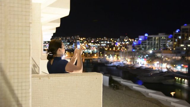a woman stands on the balcony of the hotel and takes video a nigth city on the phone