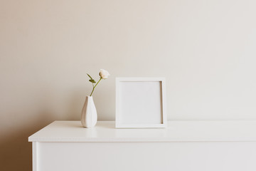 Close up of single rose in small white vase next to blank square picture frame on sideboard against...