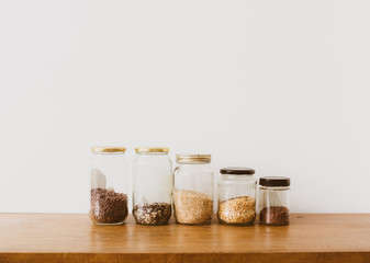 Close up of glass jars with rice, split peas and flaxseeds on oak wooden shelf against white wall -...