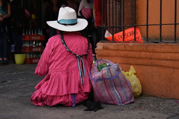 Mujer indígena con trenzas y sombrero vestida de color rosa sentada en el suelo, es una vendedora...