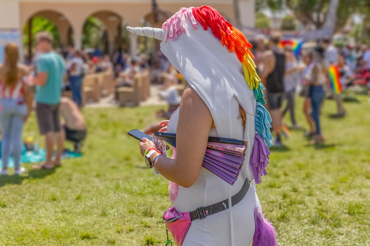 A woman in a full body unicorn costume is texting during pride. She stands off to the side facing the stage at a pride festival looking at her device. - Powered by Adobe
