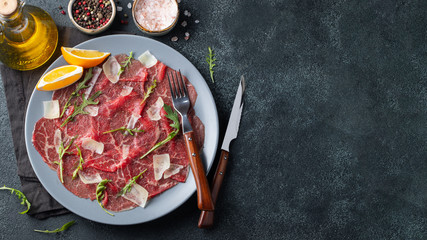 Marbled beef carpaccio with arugula, lemon and parmesan cheese on dark concrete table. Top view,...
