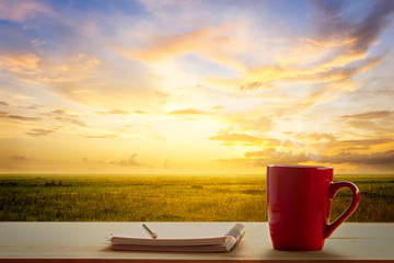 a red cup of coffee and note pad on wooden table and Empty grassland and sky at evening time