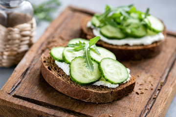 Rye bread with cream cheese and cucumber on wooden cutting board. Closeup view, selective focus, horizontal orientation - Powered by Adobe