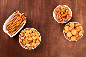 Salt crackers, sticks, pretzels, and goldfishes, shot from the top on a dark wooden background with copy space. Party snacks mix, forming a frame with a place for text