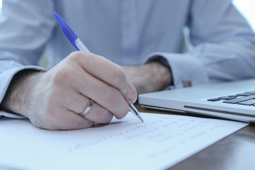 A man writes with a pen on a sheet of paper, visible computer.