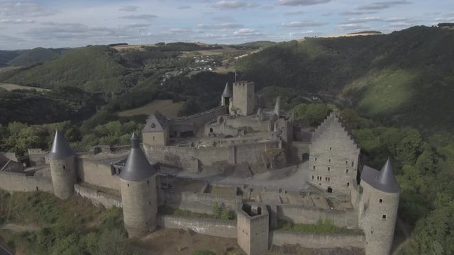 Moving above the Bourscheid castle in Luxembourg, from the back to the front. Showing the mountains, forest, village, river and bridge in the back. Sunny weather with clouds.
