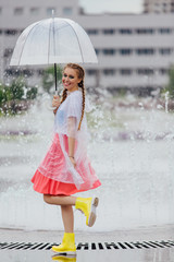 Young pretty girl with two braids in yellow boots and with transparent umbrella stands near fountain.