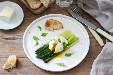 Breakfast consisting of poached egg and boiled asparagus with butter and lettuce on a wooden surface. Rustic style.