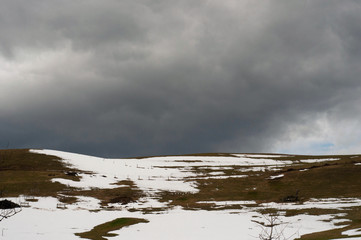 A countryside view of a hill with green grass covered with snow in early April