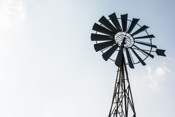 Low angle view of an old-fashioned, multi-bladed, metal wind pump atop a lattice tower in backlight against a pale blue sky.