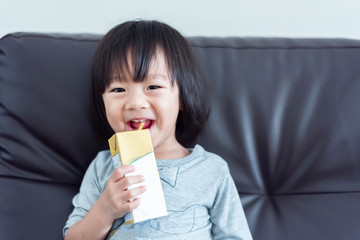 Happy Sweet Asian baby child drinking a carton of milk from box with straw on sofa