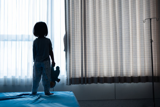Little baby child sitting on bed playing with the doll in the dark room with light throught curtain