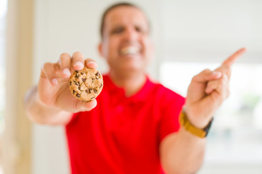 Middle age man eating chocolate chips cookies at home very happy pointing with hand and finger to the side