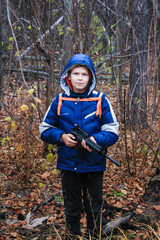 Teen boy stands in a forest in a warm jacket, holding a toy gun