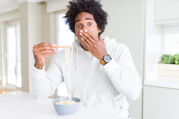 Fototapeta na wymiar African American man eating asian noodles using chopsticks at home cover mouth with hand shocked with shame for mistake, expression of fear, scared in silence, secret concept