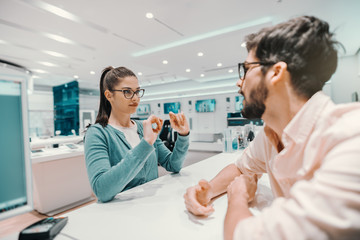 Fototapeta premium Young Caucasian charming brunette with ponytail and eyeglasses dressed casual explaining to seller which smart phone she want to buy. Tech store interior.