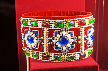 The royal crown made of gold and decorated with multi-colored precious stones stands on a stand of red velvet