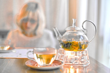 Transparent teapot with green tea on a stand with a candle on the background of a blurred breakfast blonde