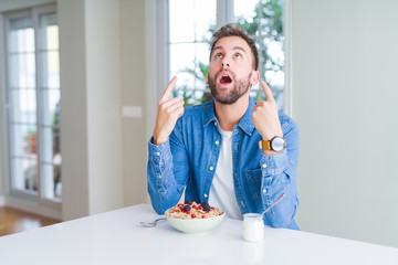 Handsome man eating cereals for breakfast at home amazed and surprised looking up and pointing with fingers and raised arms.