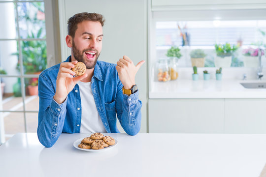 Handsome man eating chocolate chips cookies pointing and showing with thumb up to the side with happy face smiling