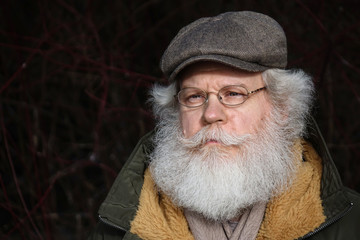 Stylish senior caucasian man wearing eyeglasses and kepi against forest background. Close up face portrait of elderly hipster with splendid mustache and beard.