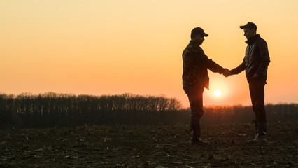 Two male farmers shake hands with each other. Stand on the background of a plowed field at sunset