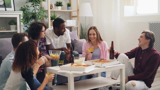 Happy men and women are talking eating and drinking clanging glasses at house party in apartment. Young people are relaxing at home together celebrating.