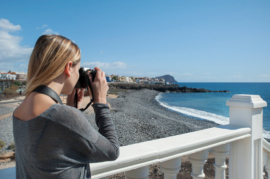 Young millennial female of Caucasian ethnicity, a remote photographer or a solo tourist, holding a digital, professional camera and taking pictures of the ocean and the small pebble beach behind