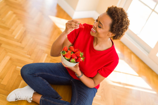 Beautiful young african woman with afro hair eating fresh strawberries sitting on the floor