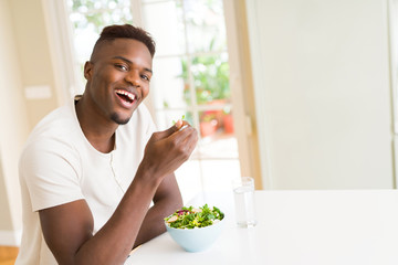 Handsome african young man eating a healthy vegetable salad using a fork to eat lettuce, happy and smiling sitting on the table