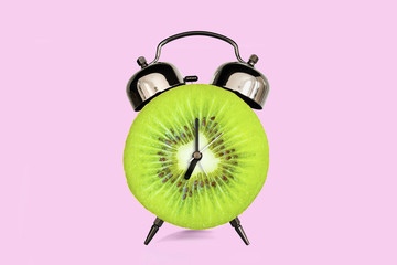 Kiwi slice on alarm clock, pink pastel background. fruit and vitamins diet at breakfast nutrition...