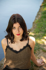 Half-length portrait of a beautiful brunette girl with red lips, sitting along a stream. Woman with long hair wearing a tank top with lace insert. Beautiful face illuminated with diffused light.