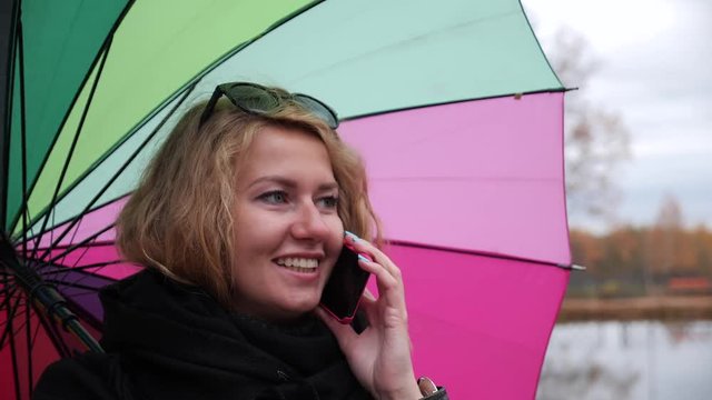 Young long-haired blonde girl with a large colorful umbrella talking on the phone in the autumn park with a smile.