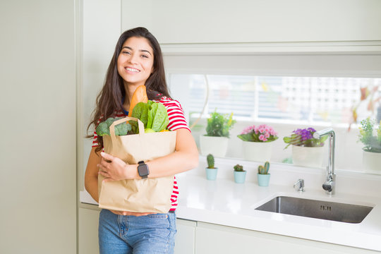 Beautiful young woman smiling holding a paper bag full of groceries at the kitchen