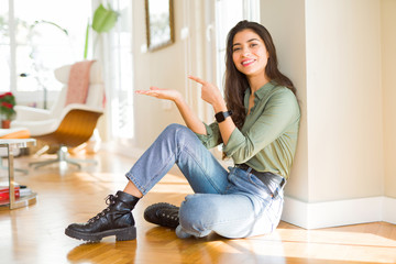 Young beautiful woman sitting on the floor at home amazed and smiling to the camera while presenting with hand and pointing with finger.