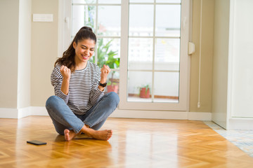 Young beautiful woman sitting on the floor at home very happy and excited doing winner gesture with arms raised, smiling and screaming for success. Celebration concept.