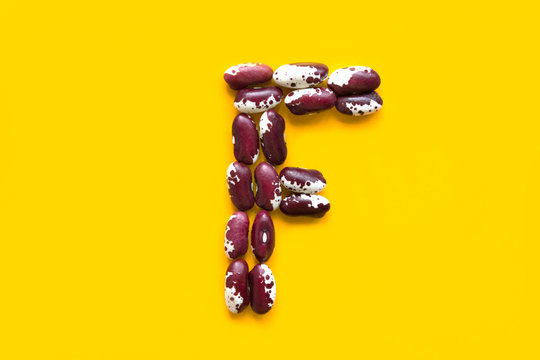 Capital Letter F From Dry Uncooked Red White Speckled Beans Legumes On Yellow Background. Creative Food Typography Art Poster. Symbol For Folic Acid. Health Plant Based Meatless Diet Wholefoods Concep