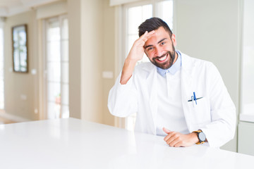 Fototapeta premium Handsome hispanic doctor or therapist man wearing medical coat at the clinic very happy and smiling looking far away with hand over head. Searching concept.