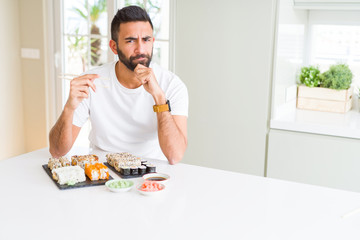 Handsome hispanic man eating asian sushi using chopsticks serious face thinking about question, very confused idea