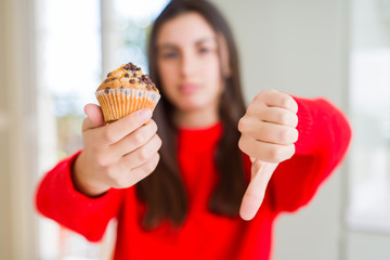 Beautiful young woman eating chocolate chips muffin with angry face, negative sign showing dislike with thumbs down, rejection concept