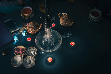 Hookah on glass table top view. cups candle and smartphone eastern tea ceremony. Stylish oriental shisha in dark with backlight. Selective focus, sugar cubes with different flavors. Copy space