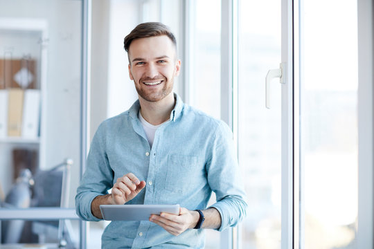 Waist up portrait of handsome young man holding digital tablet and smiling happily at camera standing by window, copy space